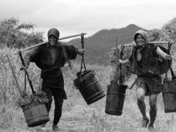 Zwei Männer tragen Eimer mit Exkrementen - Szene aus dem Film OKIKU AND THE WORLD